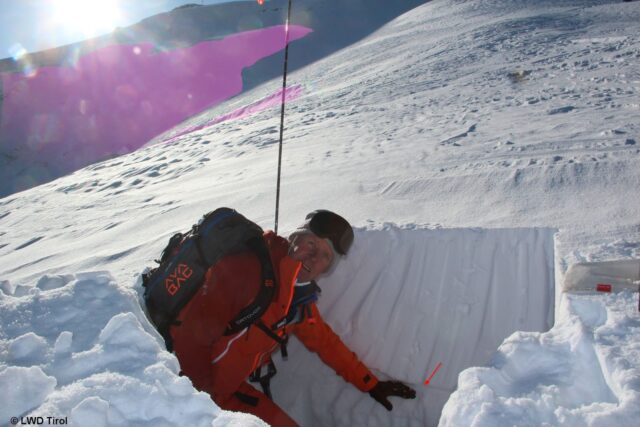Schneeprofil im Bereich des flacheren Rückens, der Richtung Gipfel zieht. Der Pfeil zeigt auf die relevante Schwachschicht. Im Hintergrund ein Teil des Lawinenanrisses. (Foto: 14.02.2022)