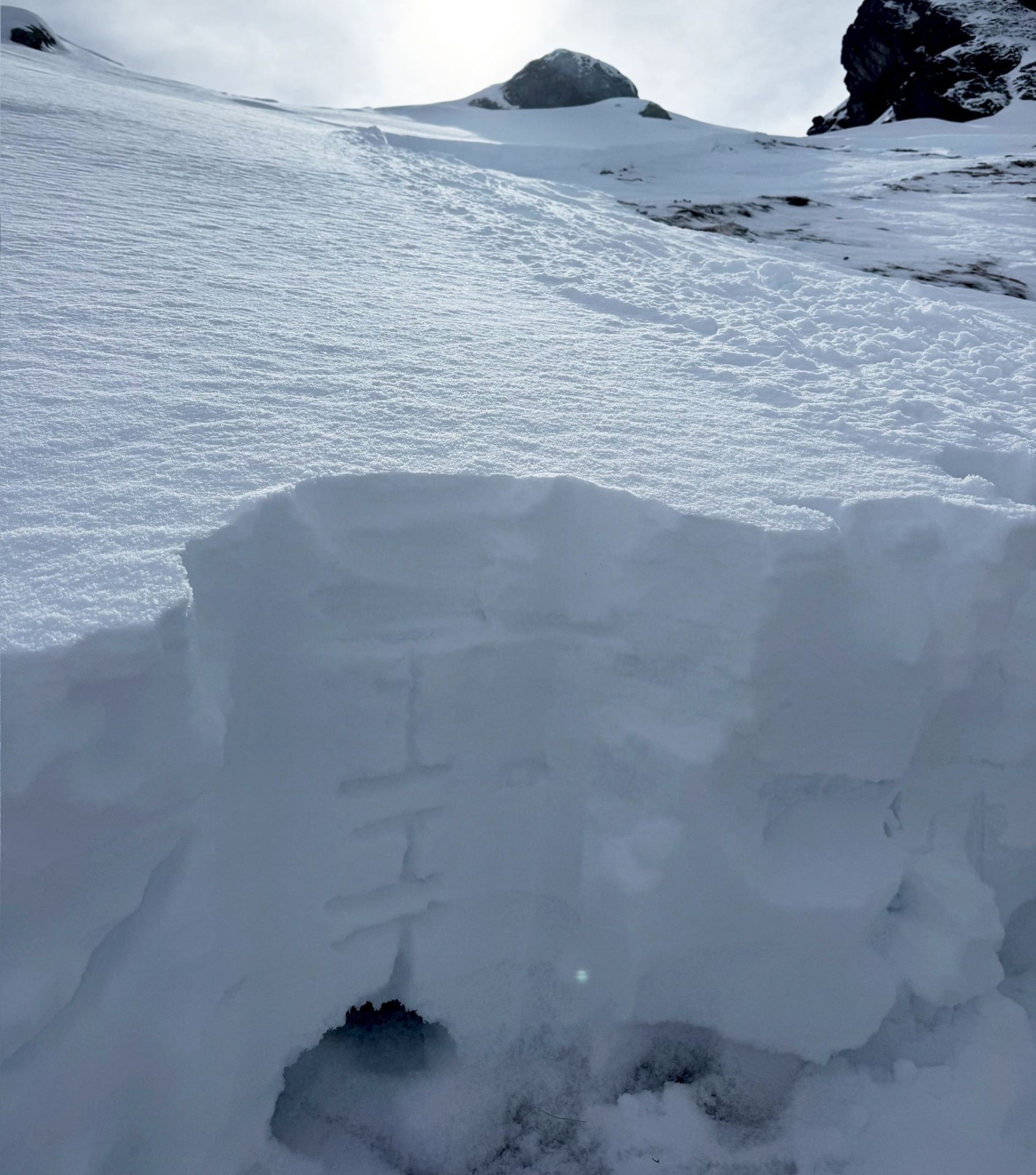 Im Hintergrund der Anriss der bereits an früherer Stelle in diesem Blog gezeigten personenausgelösten Schneebrettlawine am Pfaffenbichl an einen Nordosthang auf etwa 2300m. Im Vordergrund erkennt man im Schneeprofil die schwache Basis aus kantigen Kristallen und Tiefenreif (©Tim Sperle, 30.03.2025).